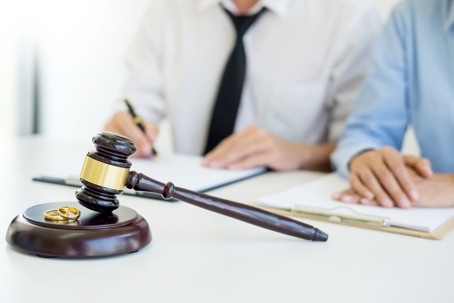 Two gold wedding bands sit on a gavel in front of a couple signing divorce papers